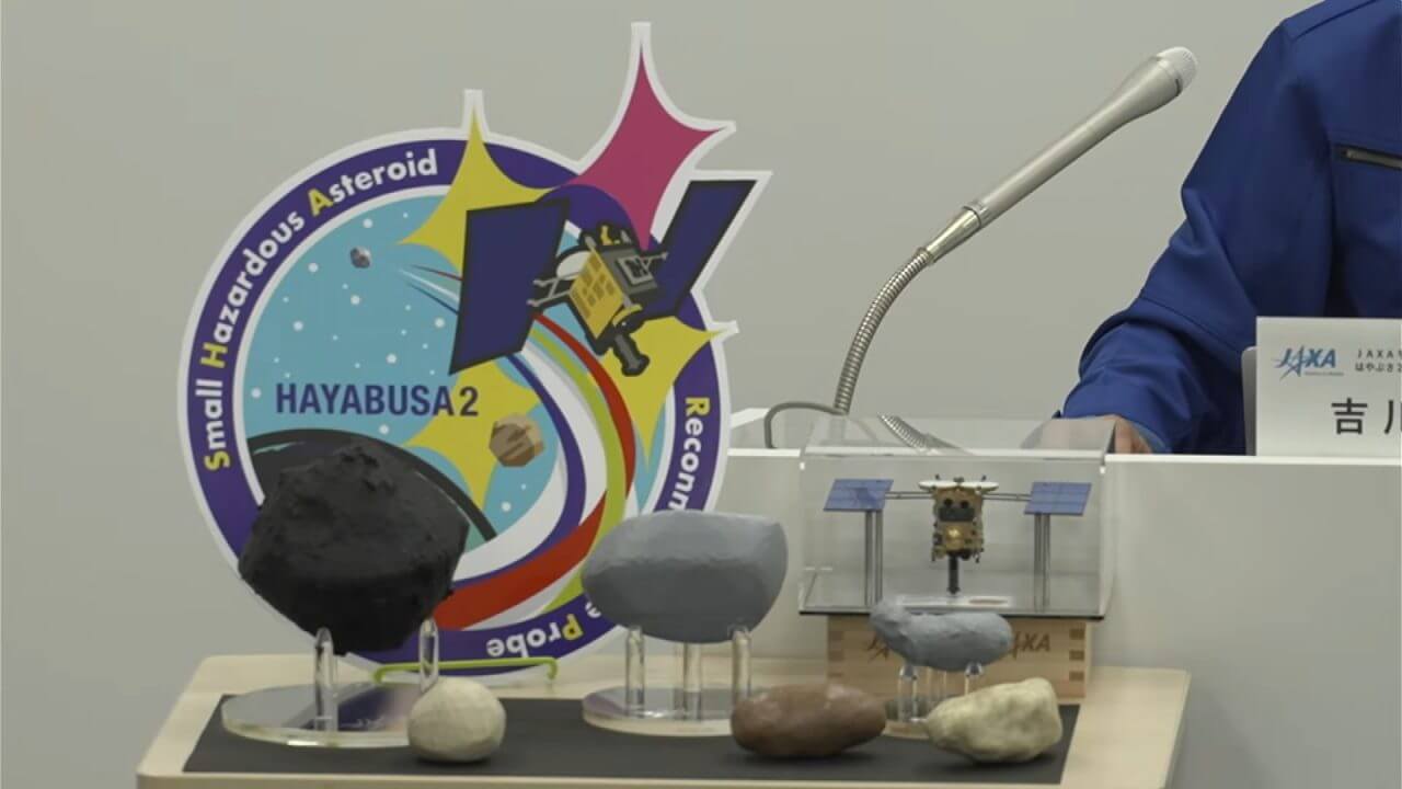 はやぶさ2拡張ミッションロゴマークと小惑星の模型。ロゴの右下に重なっているグレーの模型がトリフネで、その左隣にある黒い模型はリュウグウ。記者説明会の公式ライブ配信から引用（Credit: JAXA）